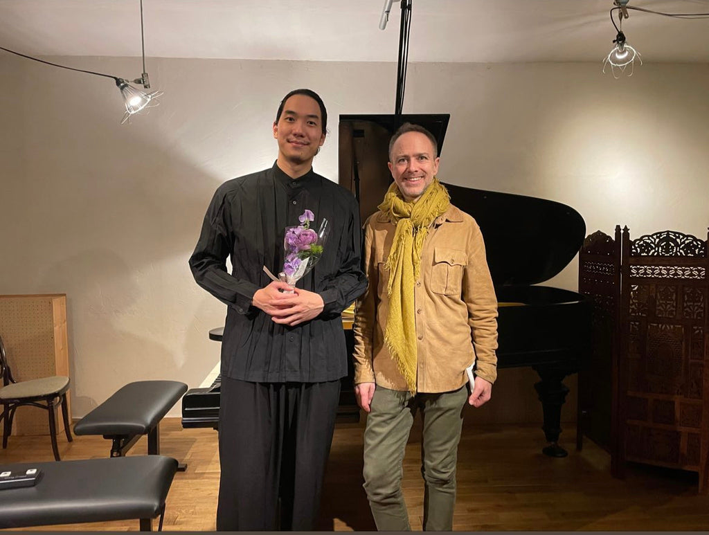 Photo de Tastsuki Sasanuma et Henri Demarquette devant un piano à queue. Henri porte une écharpe en laine j’ai e de July to June.